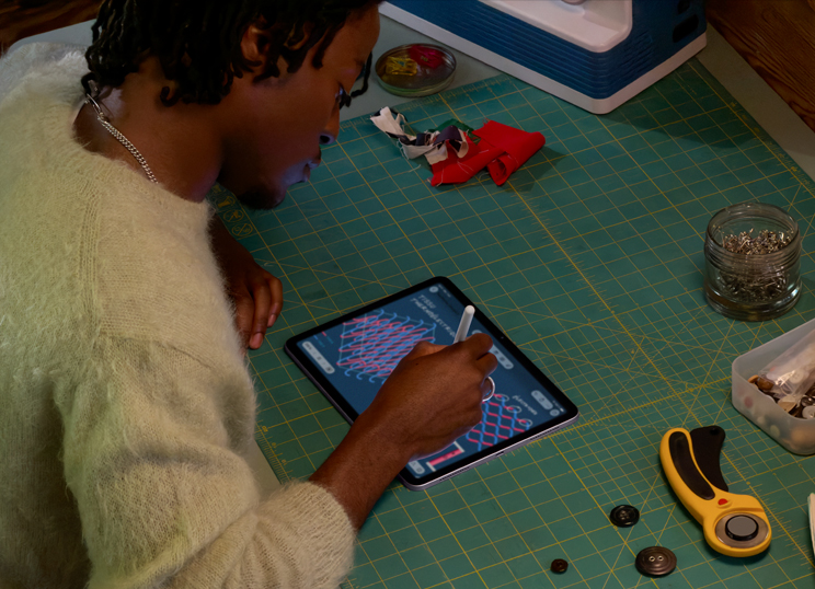Une personne dessine avec l’Apple Pencil sur un iPad posé sur un tapis de coupe vert où se trouve du matériel de couture.