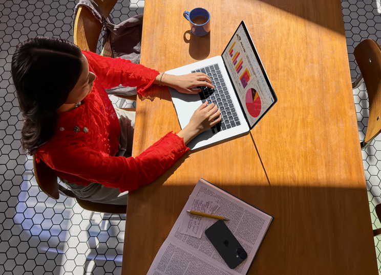 Une personne utilisant un MacBook à une table, avec une tasse, un smartphone et des papiers à proximité.