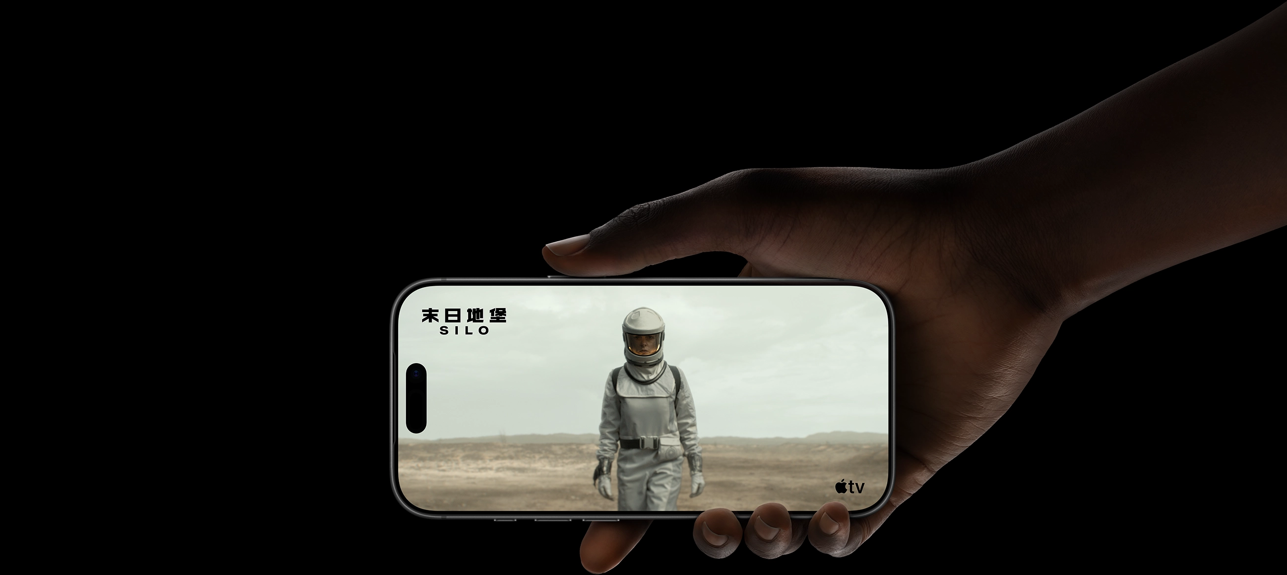 一隻手拿著 iPhone 17 Pro，螢幕上顯示一名身穿防護服、頭戴頭盔的女子，走過一片貧瘠的景觀。Apple TV 標誌，劇集標題：《末日地堡》(Silo) 