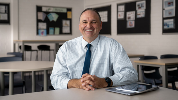 Le directeur Mark Watkins souriant, assis à un pupitre avec un MacBook fermé et un iPad près de lui.